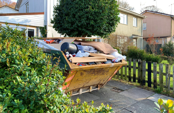 An image of a skip that is full of rubbish on a driveway surrounded by bushes.