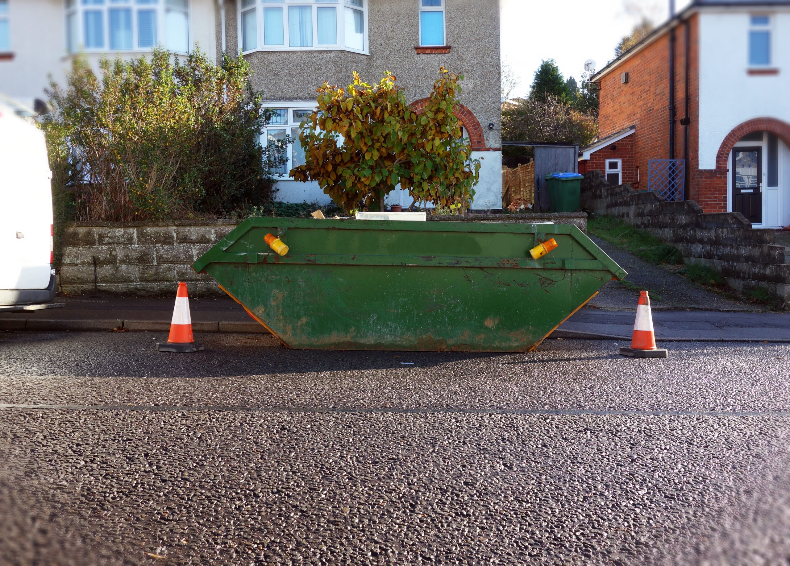 A green skip located on the road with orange safety cones around it, requiring a skip permit.